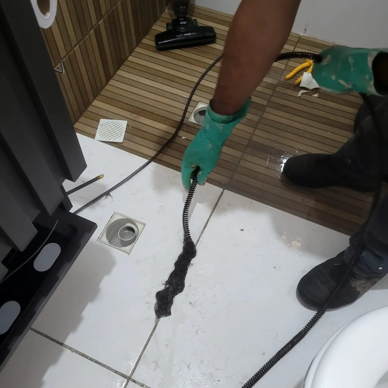 Profissional usando cabo desentupidor para retirar o resíduo que entupia o ralo de um banheiro.
